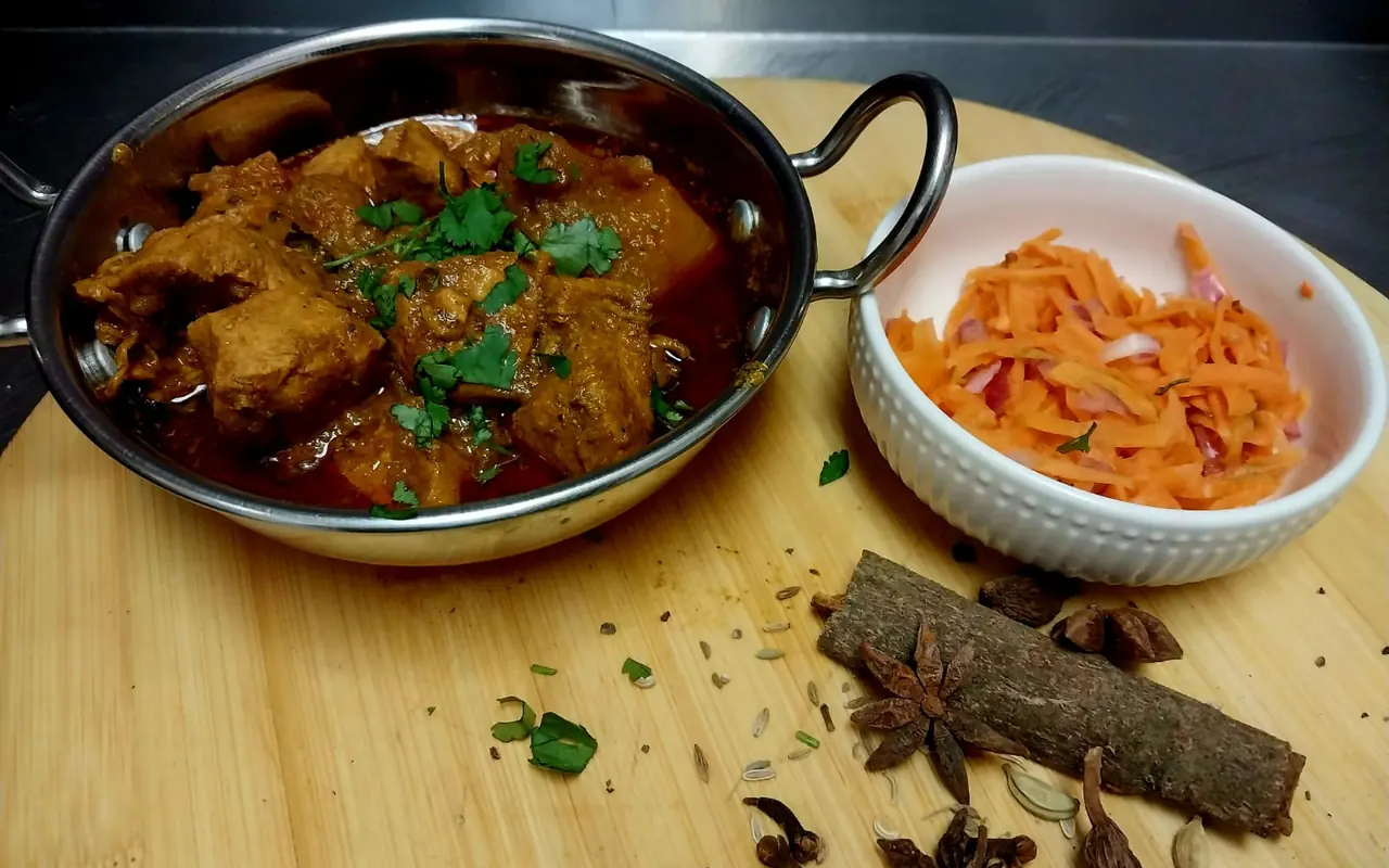 Chicken curry in a steel karahi with carrot salad and whole spices on a wooden board