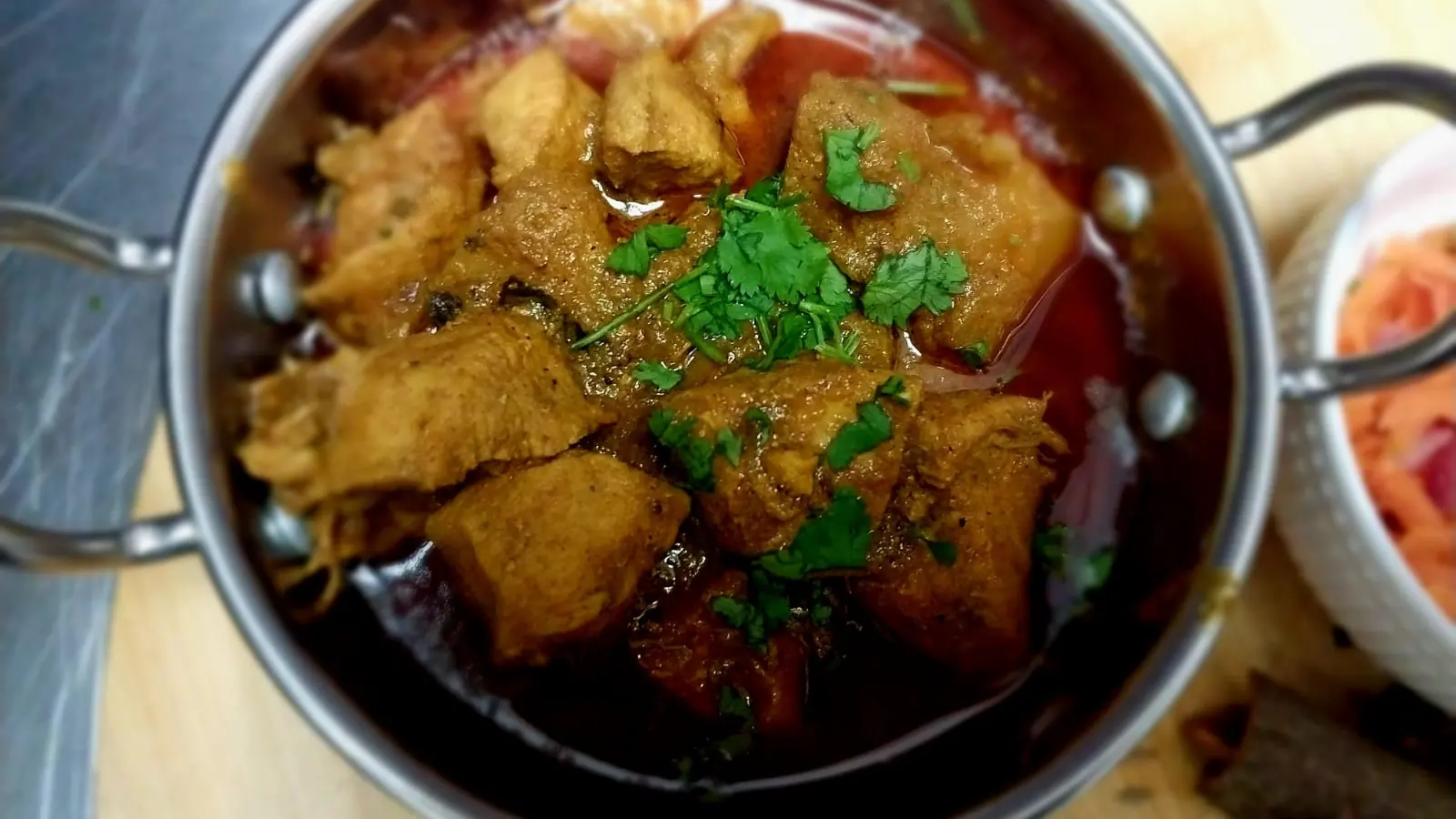 Chicken curry in a steel karahi on a wooden board with carrot salad and whole spices
